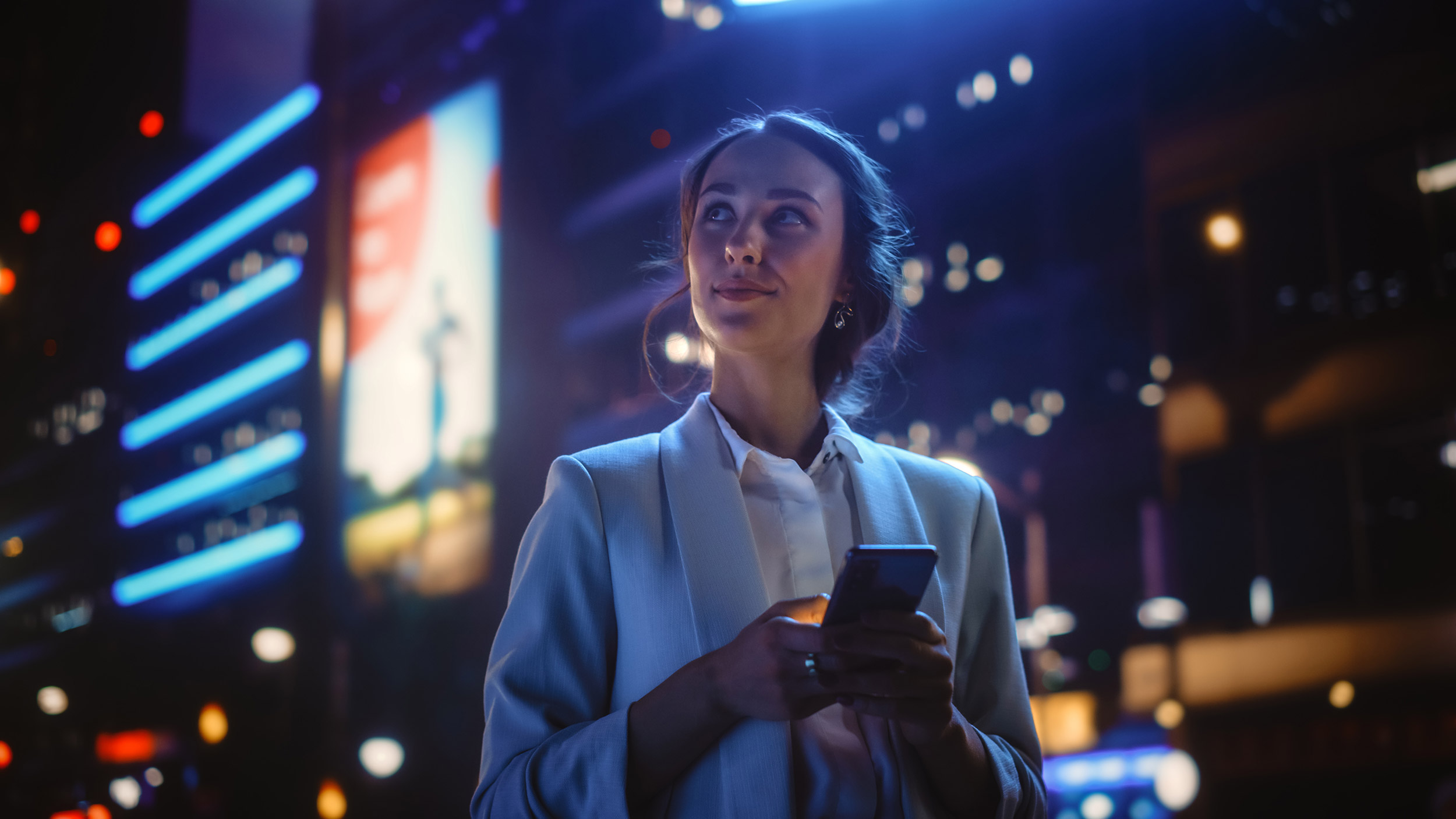 Woman in business attire holding a mobile phone in an urban environment with atmospheric lighting